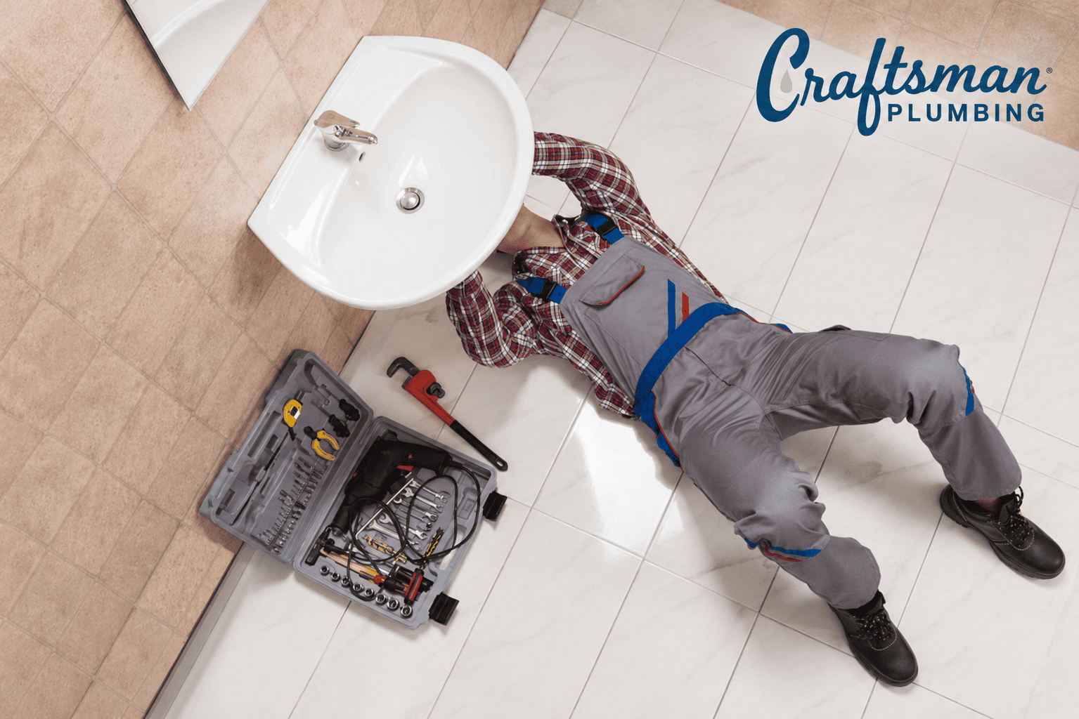Plumber lying under a bathroom sink with tools nearby, repairing plumbing beneath a wall-mounted sink, with the Craftsman Plumbing logo in the upper right corner.